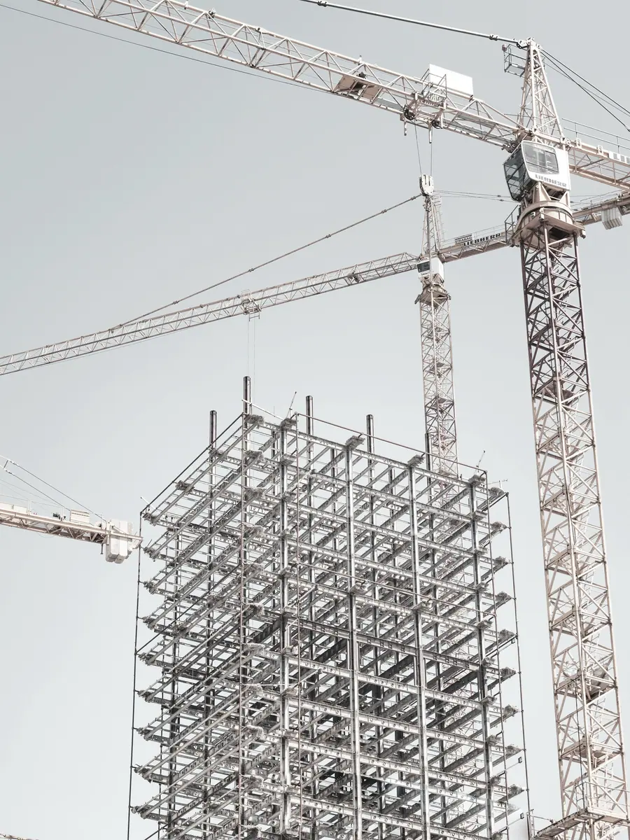 Estrutura metálica de um edifício em construção, com duas gruas ao lado. Fotografia de referência, em tom mineral.